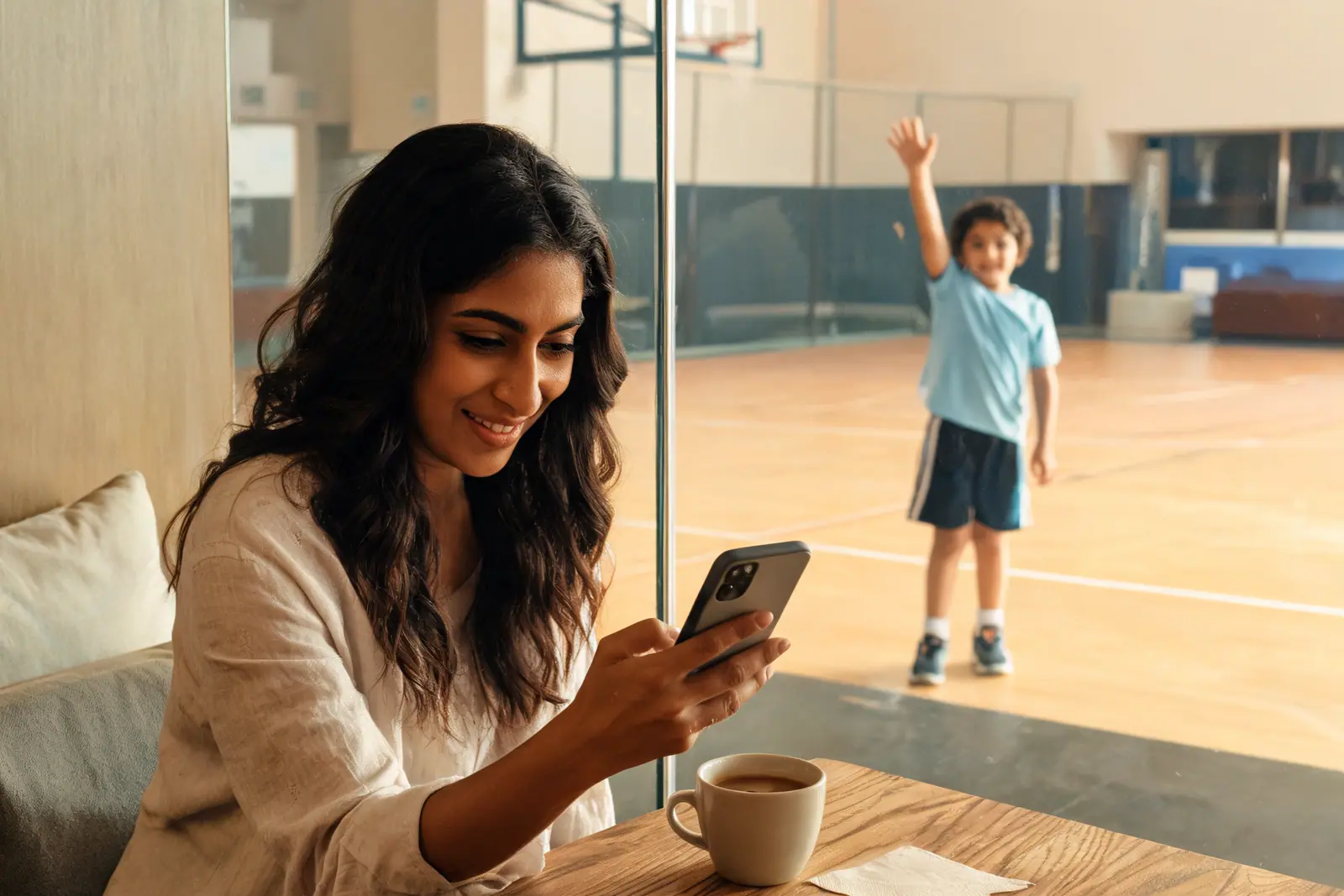 A mother smiling at her phone beside a sports hall while her son waves from the court