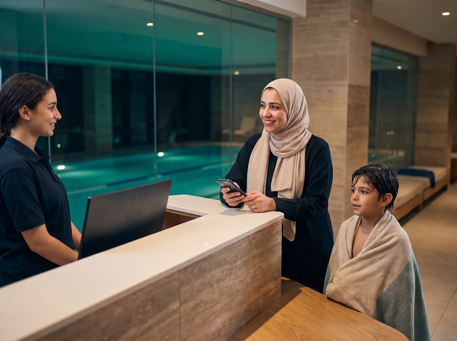 A receptionist at a pool's front desk checking in a mother and her son, wrapped in a towel after his lesson
